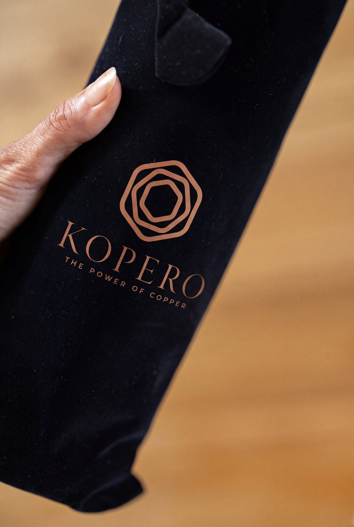 Een hand houdt de zwarte fluwelen draagzak van Kopero vast, met het koperkleurige logo erop