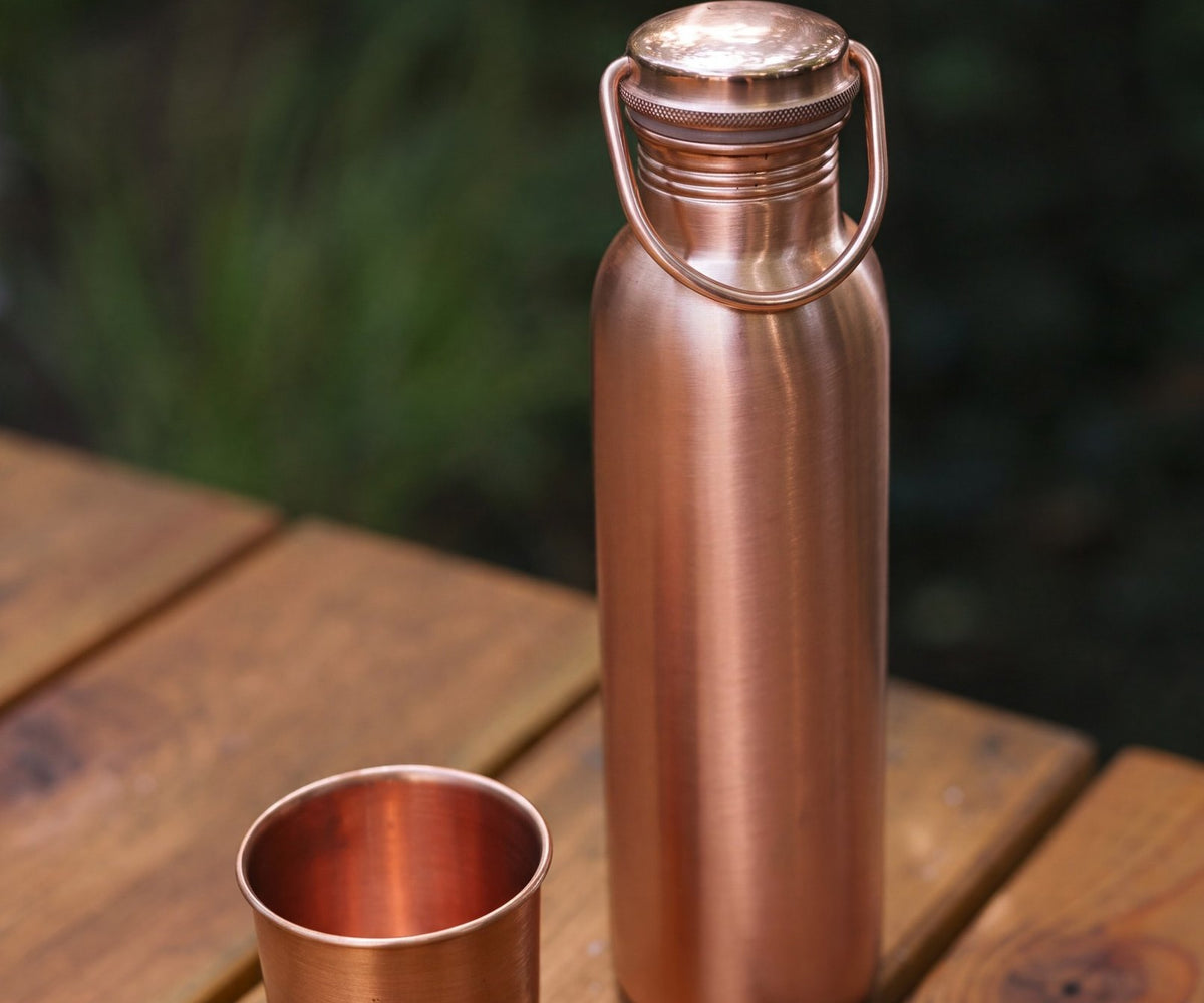 Kopero Koperen Waterfles Glad, op een houten tuintafel naast een koperen beker