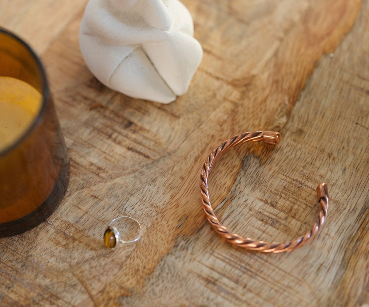 Kopero Koperen Armband Gedraaid, op een houten tafel naast een beeldje en een ring