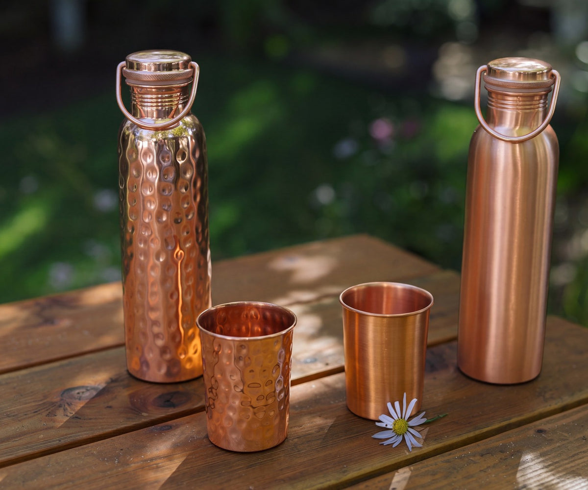Een gehamerde en een gladde koperen waterfles met twee koperen bekers op een houten tuintafel, met een madeliefje