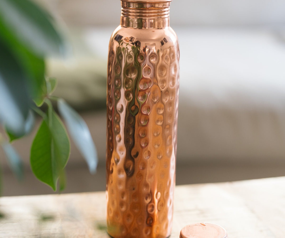 Kopero Koperen Waterfles Gehamerd, staand op een houten tafel voor een bank, met de losse dop ernaast
