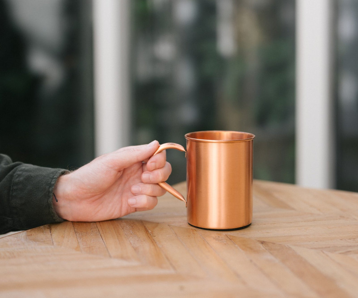 Kopero Koperen Mok Glad, op een houten tafel, met een hand aan het handvat