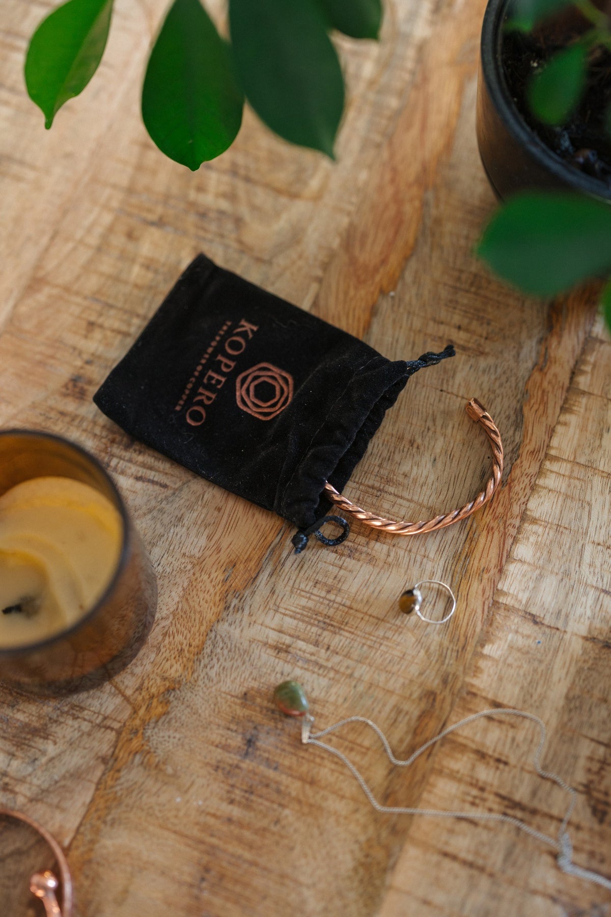 Kopero Koperen Armband Gedraaid, op een houten tafel naast de fluwelen draagzak, een kaars en sieraden