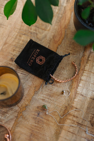 Kopero Koperen Armband Gedraaid, op een houten tafel naast de fluwelen draagzak, een kaars en sieraden