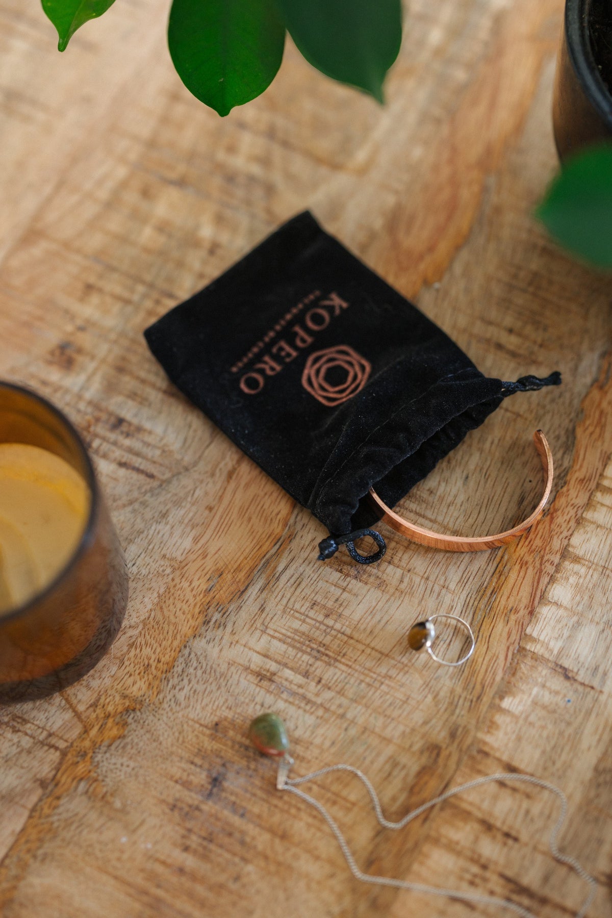 Kopero Koperen Armband Glad, op een houten tafel naast de fluwelen draagzak, een kaars en sieraden