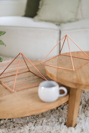Kopero Koperen Piramide Energizer Gedraaid Groot (23,56 cm), een gedraaide en een gladde grote piramide op twee houten tafels met een mok