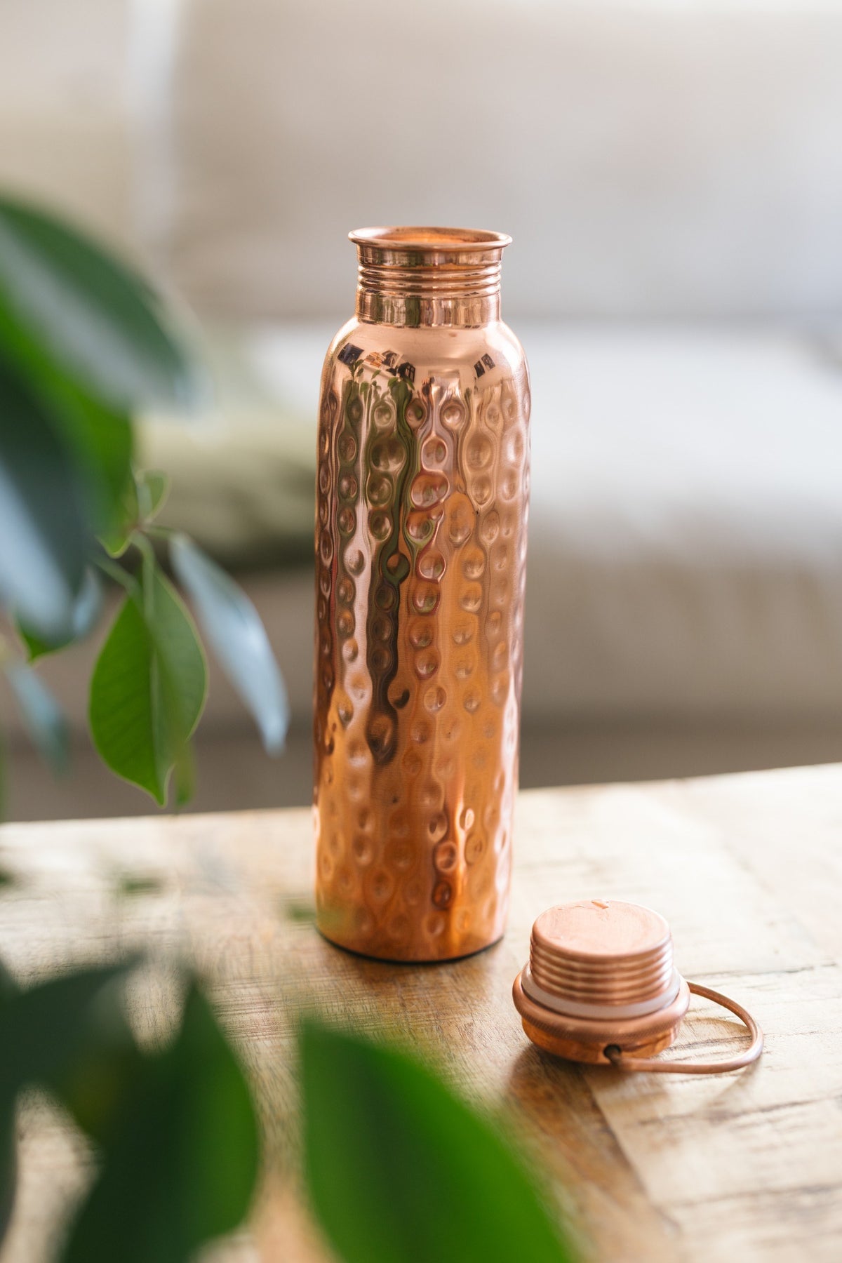 Kopero Koperen Waterfles Gehamerd, staand op een houten tafel voor een bank, met de losse dop ernaast